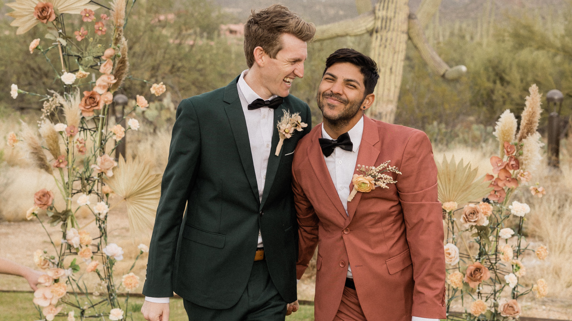 Two grooms smiling at each other during an outdoor desert wedding with neutral-toned floral decorations.