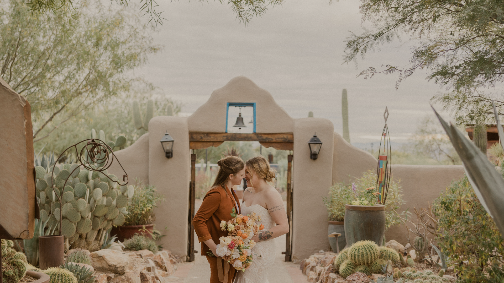 Two brides sharing a tender moment in front of an adobe-style desert venue entrance, holding a bouquet of colorful flowers.